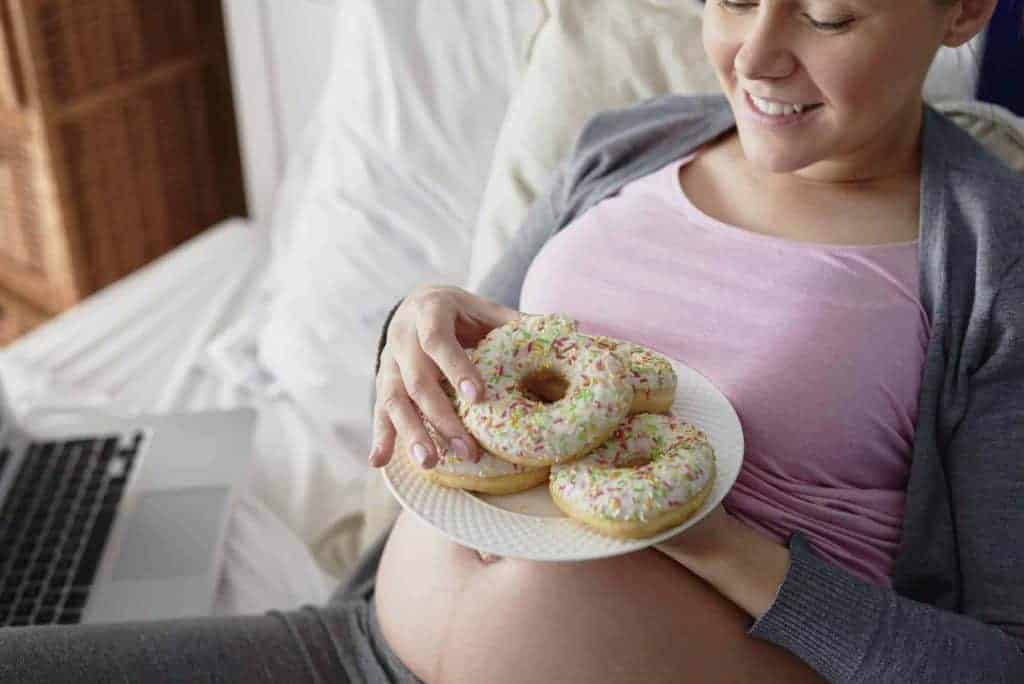 Pregnant woman eating colorful donuts in bed